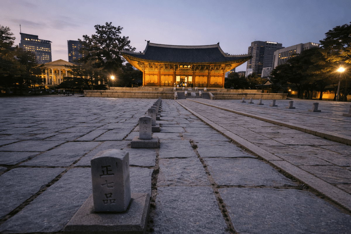 need for belonging — empty stone path leading to illuminated Korean palace at dusk, city buildings behind