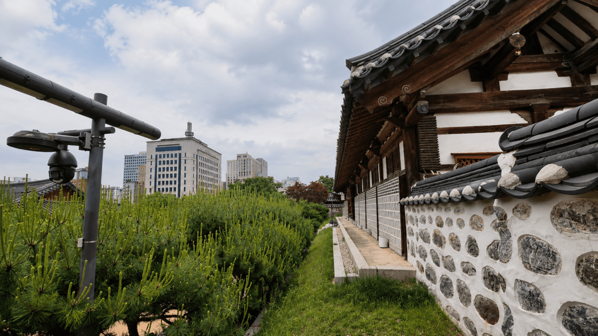 Hanok stone wall path with pine trees and modern Seoul buildings beyond — what happens when you stop pulling away accumulates
