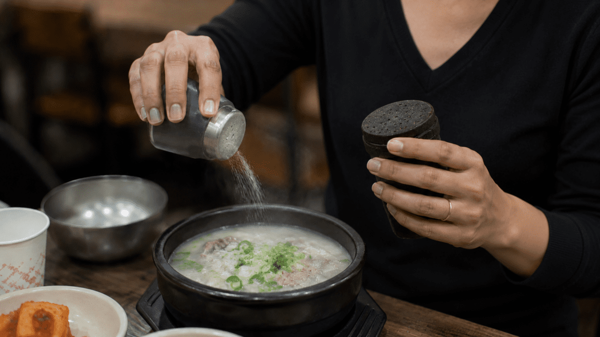 Close-up of hands seasoning a bowl of Korean bone broth with salt, green onion floating on top of the milky white soup