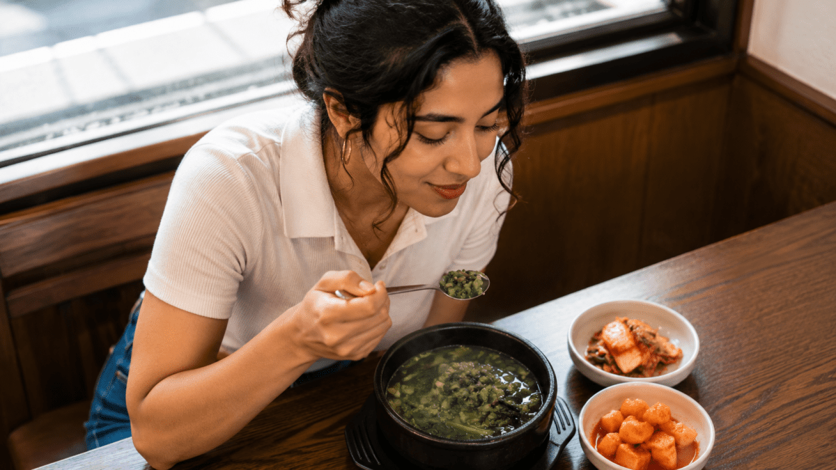 Woman lifting a spoonful of olgaengi soup with greens and shellfish, a Korean hangover soup from Chungcheong served with side dishes