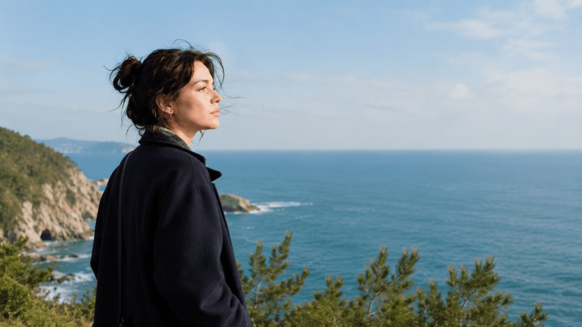 Woman standing alone on a coastal cliff, hair blowing in the wind, looking out at the sea, lonely after talking to parents