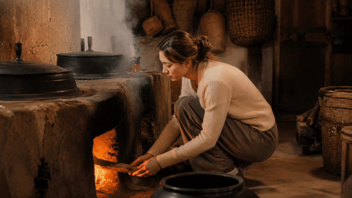 Woman feeding firewood into a traditional agungi stove, the fire behind ondol Korean floor heating's age-old warmth