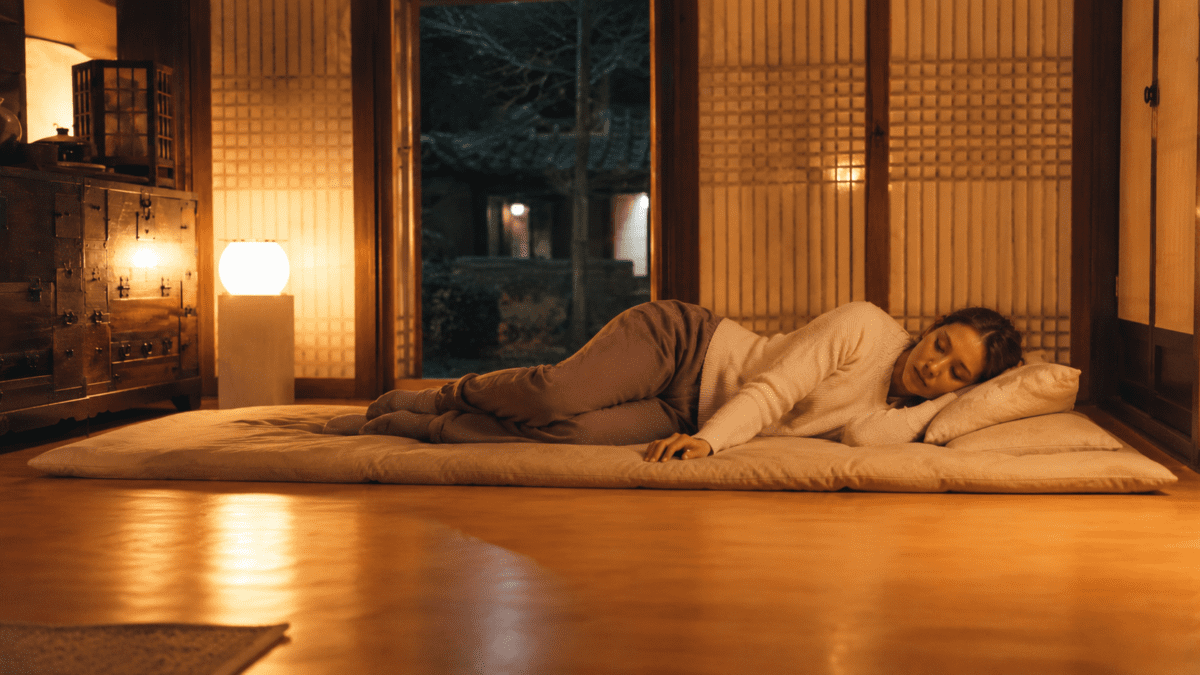 Woman sleeping peacefully on a floor mat in a warmly lit hanok room, ondol Korean floor heating keeping the night gentle