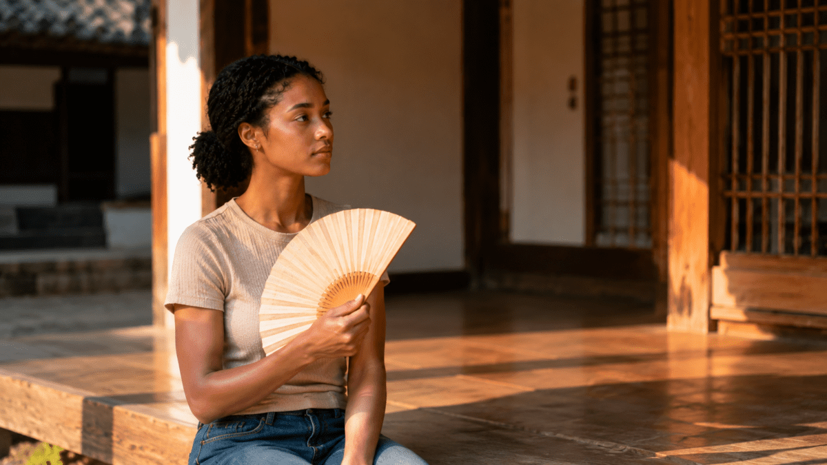 Woman fanning herself on a hanok porch in late sunlight, reflecting the lingering heat in the afternoon cycle meaning.