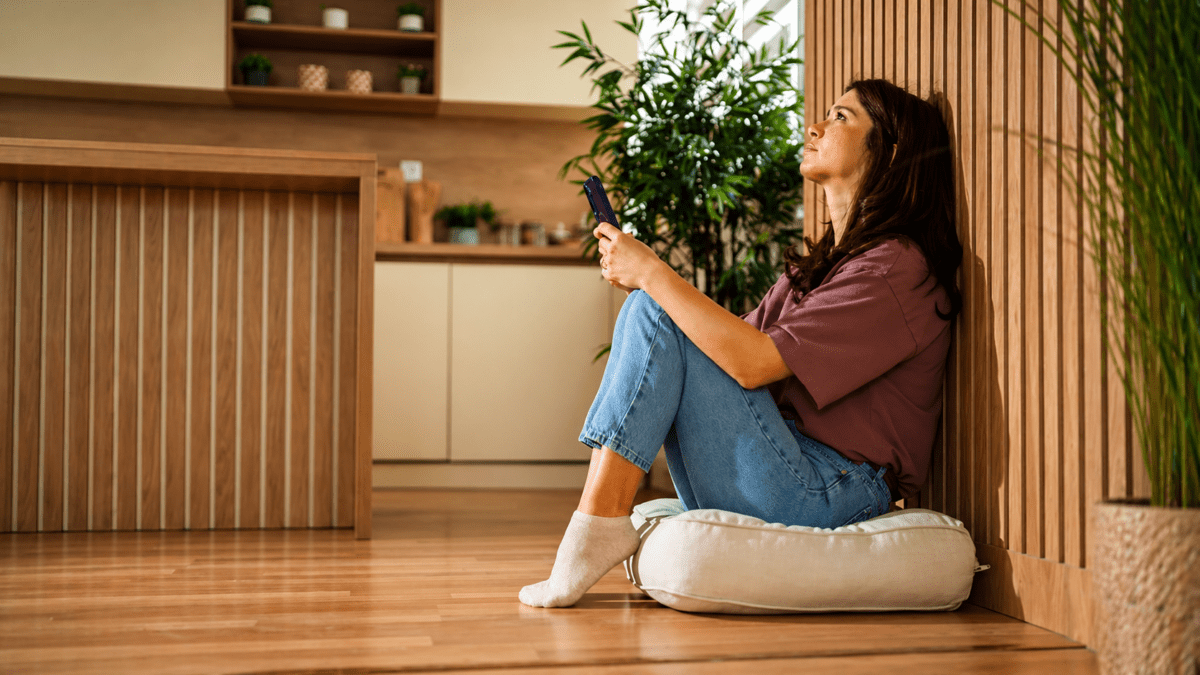 A woman sitting alone on a floor holding her phone, looking upward with anticipation and waiting, representing the anxious attachment style's experience of seeking reassurance and dreading disconnection in relationships.