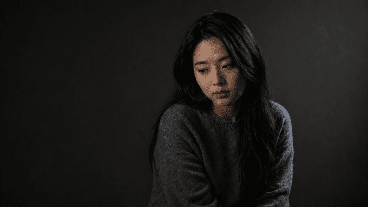A woman with a contemplative, worried expression looking downward against a dark background, representing the anxious attachment style's internal experience of uncertainty and longing for reassurance.