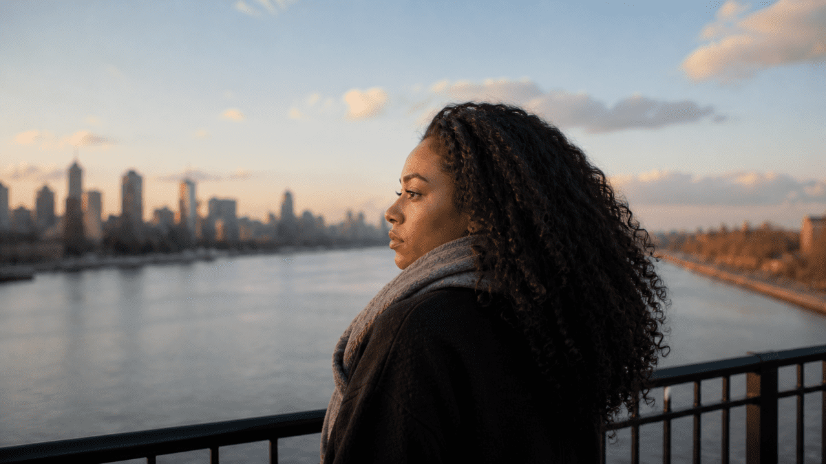A woman standing by a waterfront overlooking a city skyline, looking back over her shoulder with a peaceful yet resolute expression, representing the understanding that attachment styles are learned patterns that can shift and change through conscious awareness and safe relationships.