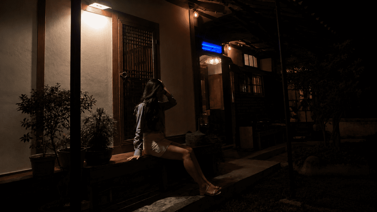 Woman standing in a hanok courtyard at night holding her phone, mid-check—the automatic ritual of looking at her balance without truly seeing it, caught between reassurance and avoidance.