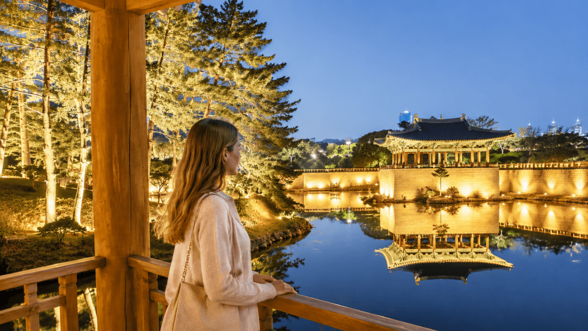 conversation replay — woman standing quietly on a wooden pavilion at night, looking across a calm river toward an illuminated Korean palace reflected in the water.