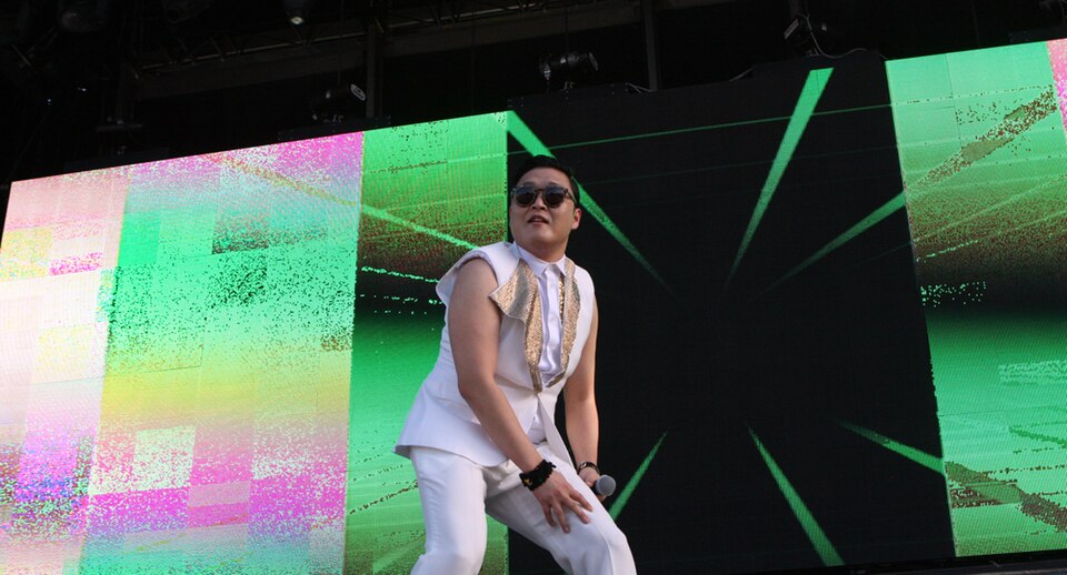 Psy performing Gangnam Style dance on stage during a live concert with bright digital visuals behind him