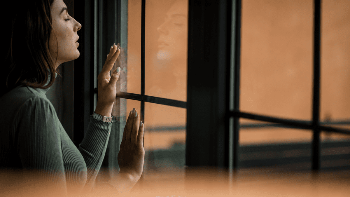 Woman looking out a window with her hand pressed against the glass, a moment of reflection on the endless cycle of goalpost shifting—where every achievement moves the target further away.