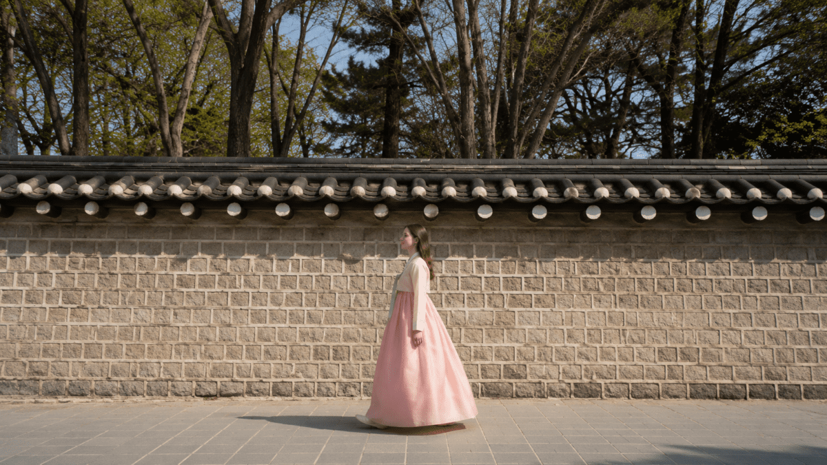Woman in hanbok walking toward Seosulla gil, passing traditional stone walls and quiet old Seoul streets on a sunny afternoon.