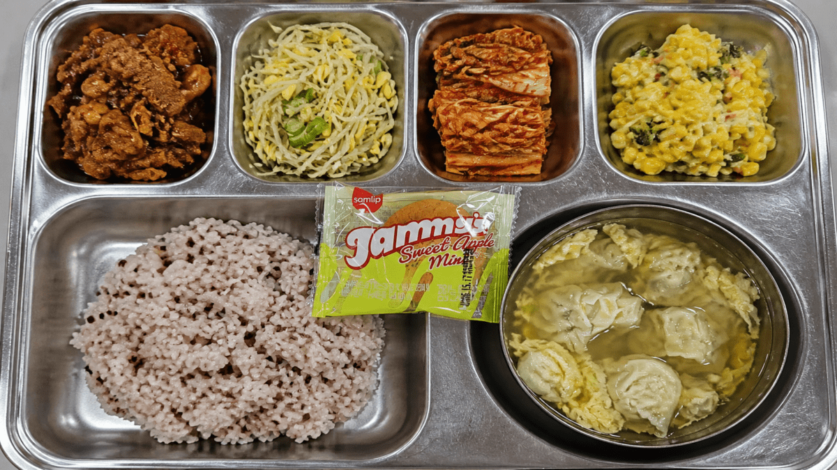A Korean school lunch tray with multigrain rice, braised meat, stir-fried noodles, seasoned vegetables, corn, and soup with an egg roll.