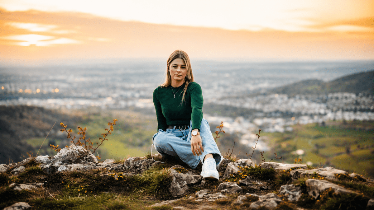 Woman sitting confidently on hilltop overlooking vast landscape at sunset, representing the freedom and self-acceptance of living life without asking permission