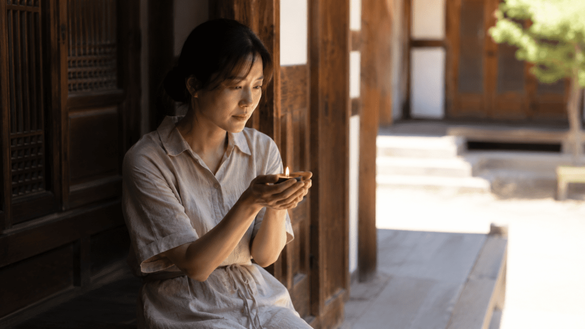 Woman cupping a small candle flame in a wooden hanok corridor, embodying the inner flame in the summer solstice meaning.