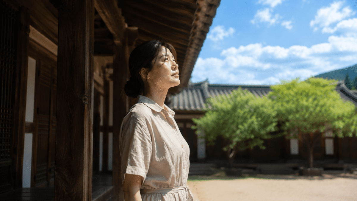Woman standing under a hanok eave looking up at a bright sky, marking the threshold between shade and peak light in O.