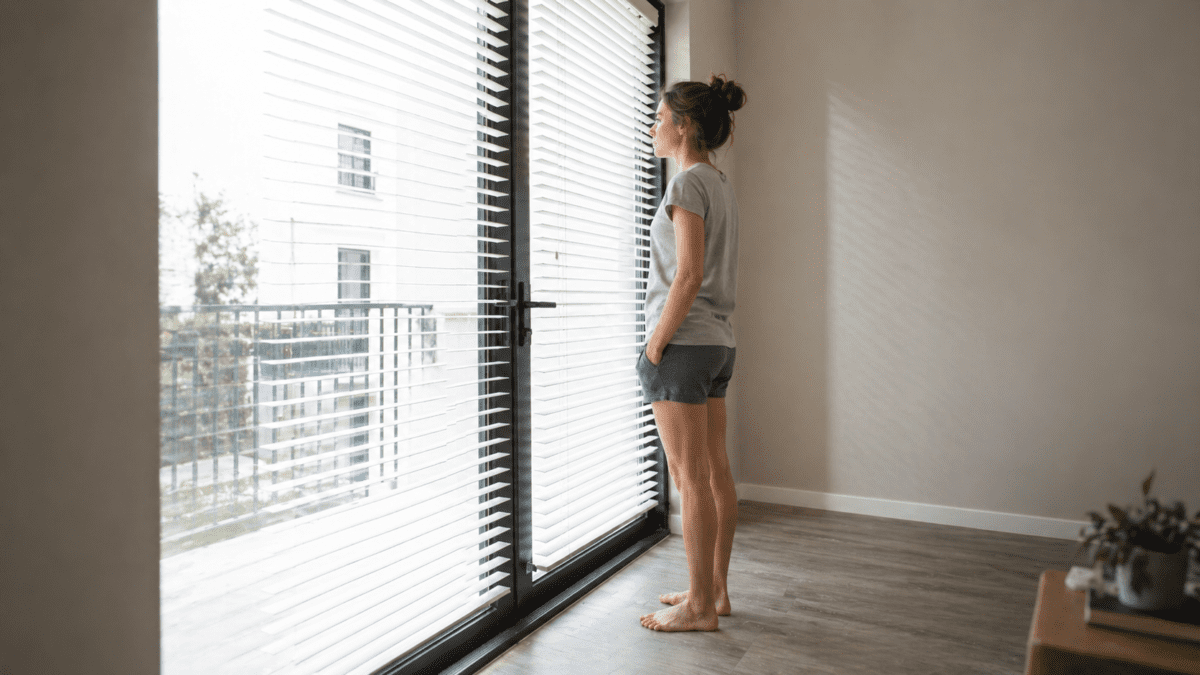 A person standing at her apartment window looking out at the city, representing temple slow food—illustrating how the rhythm dissolves back into ordinary life, but the knowledge that the gaps exist and can be left open returns her again and again.