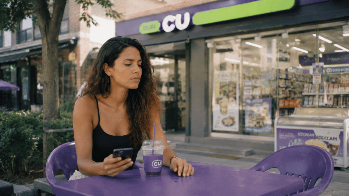 Woman sits outside a convenience store, phone in hand, drink untouched, held just far enough from tomorrow to feel safe.