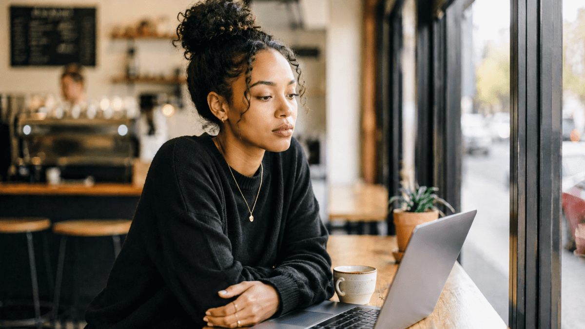 Woman sits at a café window, laptop open but untouched, the quiet pause before changing routines that no longer fit.