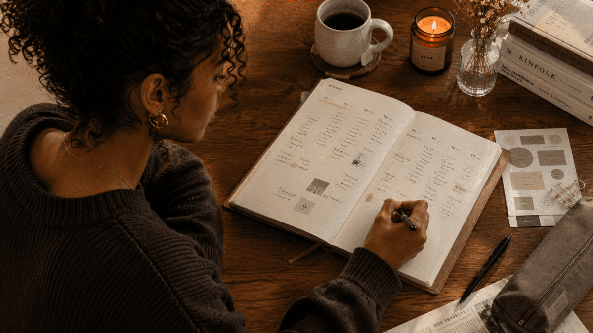 Woman writes in her Korean planner weekly spread at a warm desk, with a coffee mug, candle, and books arranged beside the open planner.