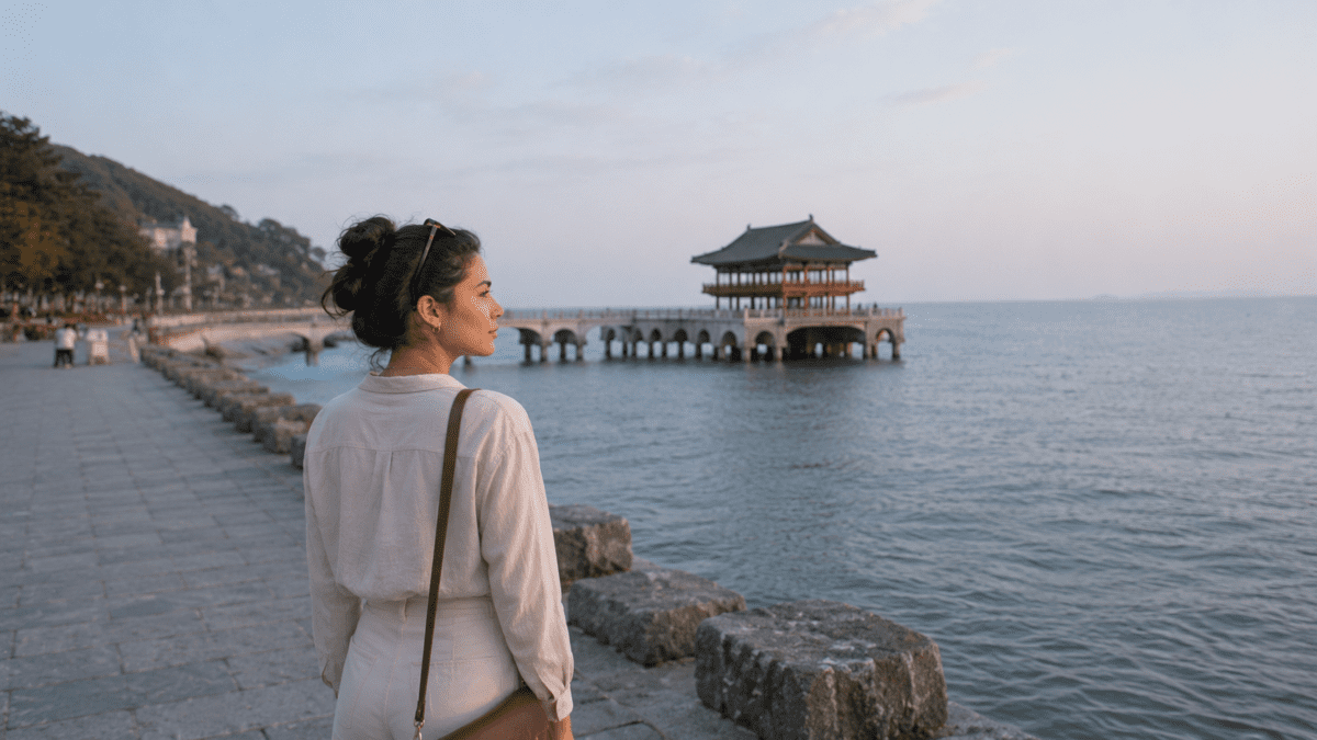 A woman stands still by the sea at dusk, the day performed well and now gone — people pleasing exhaustion settling in the quiet after.