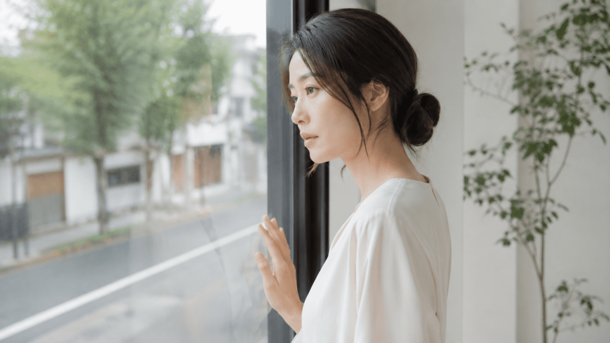 A woman stands at a window, one hand on the glass, watching the street below — seeing the people pleasing pattern clearly for the first time.