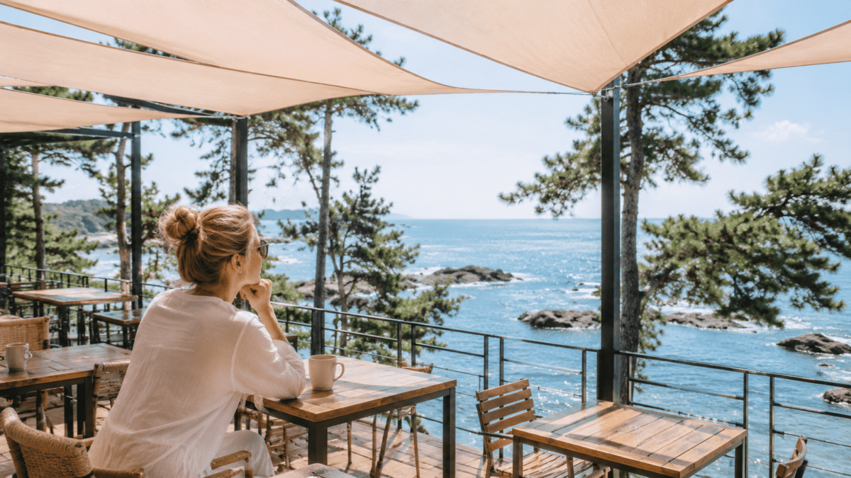 A woman sits at a Busan café terrace under a shade sail, where pure support frames the view without closing it off.