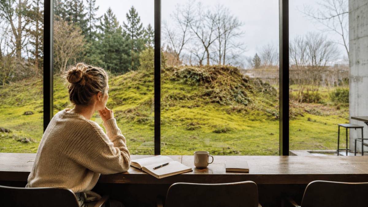 A woman sits with an open notebook and coffee at a forest-view Korean café — the Korean café experience where permission to simply exist comes with the seat.