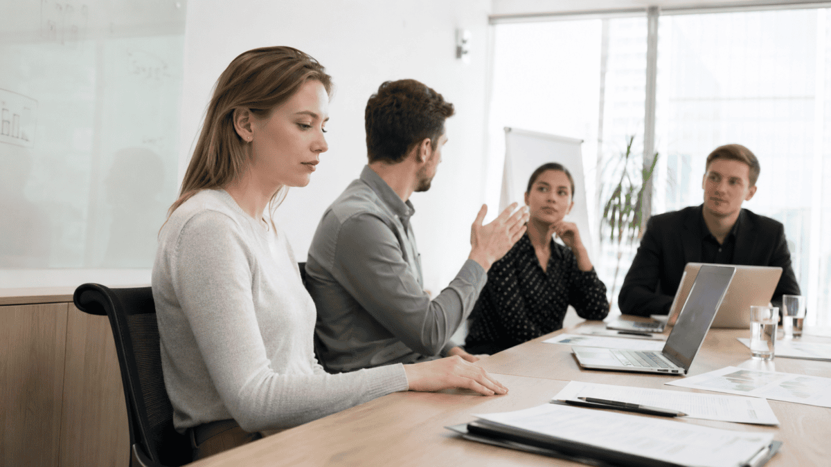 A young white woman sits quietly at a meeting table with her hands down while a male colleague volunteers for a task, illustrating quiet quitting in the workplace.