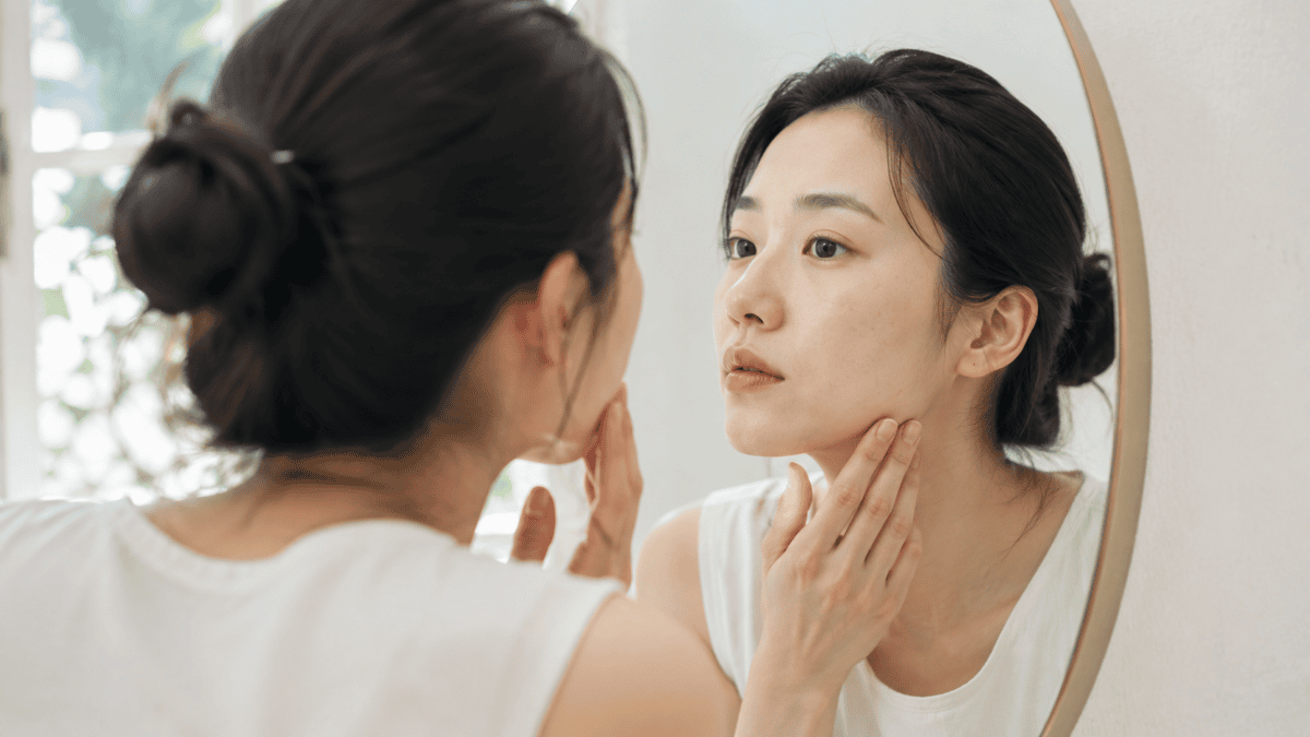 A woman studies her own face closely in the mirror, seeing the subtle aesthetic that others only sense as looking well.