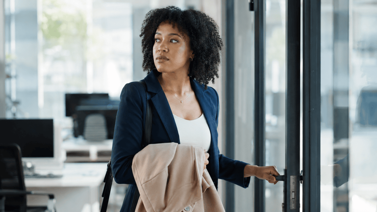Woman pauses at the office doorway, coat in hand, looking back at her desk — the sunk cost fallacy of not leaving.
